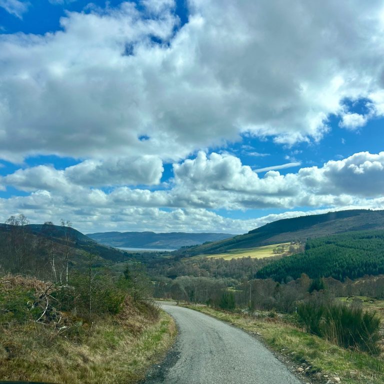 Looking over Drumnadrochit from Beauly side looking from the hills over Drumnadrochit with Loch Ness in the background. The sky is blue and the clouds are white.